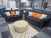 An elegant black Chesterfield sofa with button-tufted design, scroll arms, and metallic-studded detailing, accompanied by plush orange and white pillows, placed in a showroom with modern decor.